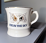 A white mug with a gold-sketch of an owl's upper head and underneath the slogan "Eye in the Sky"